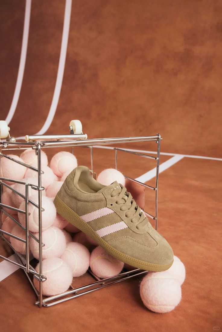 Olive suede sneaker on chrome basket of pink tennis balls on terracotta court