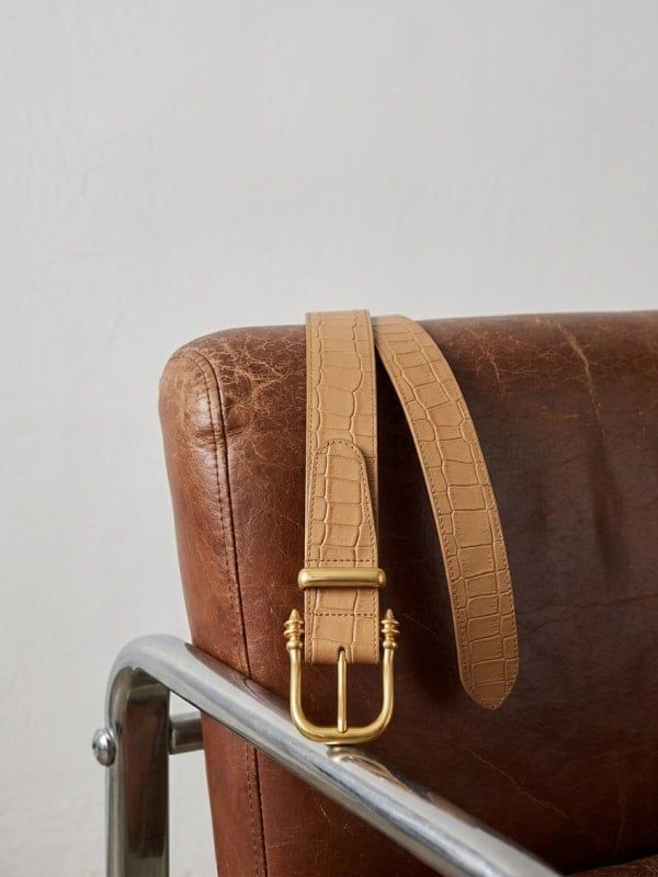Tan croc belt looped over the chrome arm of a vintage brown leather lounge chair, in soft natural light.