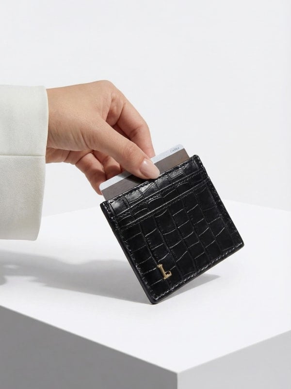 Cropped shot of a hand in a white blazer cuff sliding a credit card into a black croc-embossed card holder placed on a clean white pedestal.