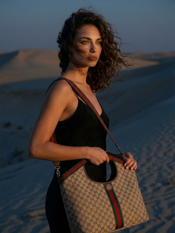 Woman with curly dark hair in a black bodysuit carrying a Gucci GG monogram hobo bag, walking through golden sand dunes at sunset.