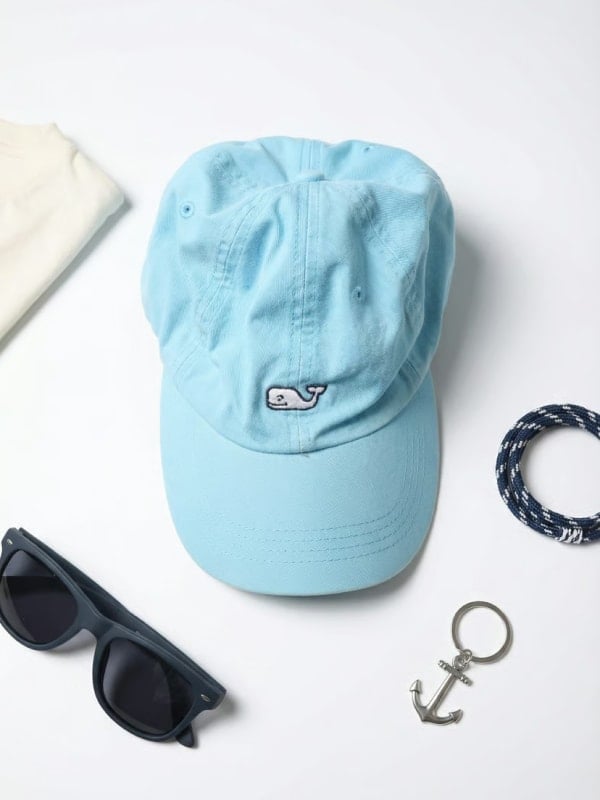 Light blue whale-logo cap flat-lay styled with black sunglasses, a folded cream sweatshirt, and an anchor keychain on a clean white background.