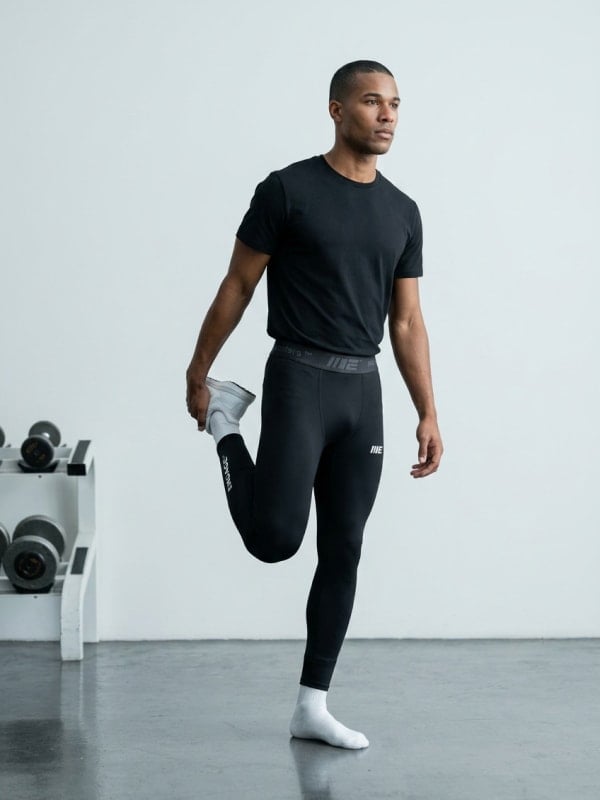 Man in a black t-shirt and Engage compression tights stretching one leg in a sunlit studio gym, with a dumbbell rack in the background.