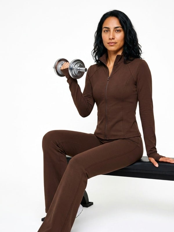 Woman in a brown mock-neck zip jacket and flared activewear pants seated on a weight bench, lifting a single dumbbell against a clean studio backdrop.