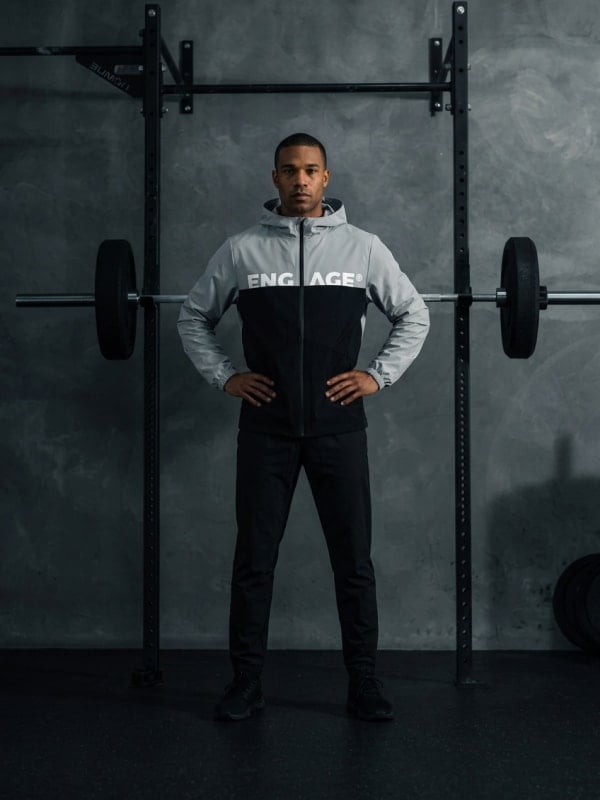 Man in a grey and black Engage zip-up windbreaker standing in front of a barbell rack inside a concrete-walled gym.