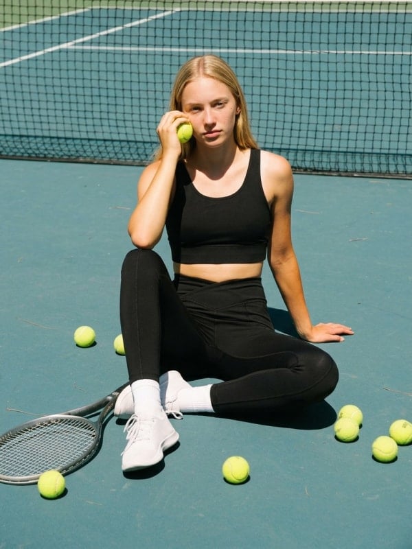 Blond woman seated on a tennis court in a black sports bra and leggings, holding a tennis ball with a racket and scattered tennis balls around her.
