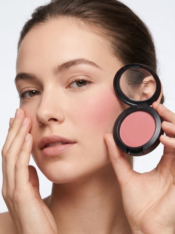 Brunette woman with glowing skin holding an open MAC blush compact next to her cheek in a clean studio setting.