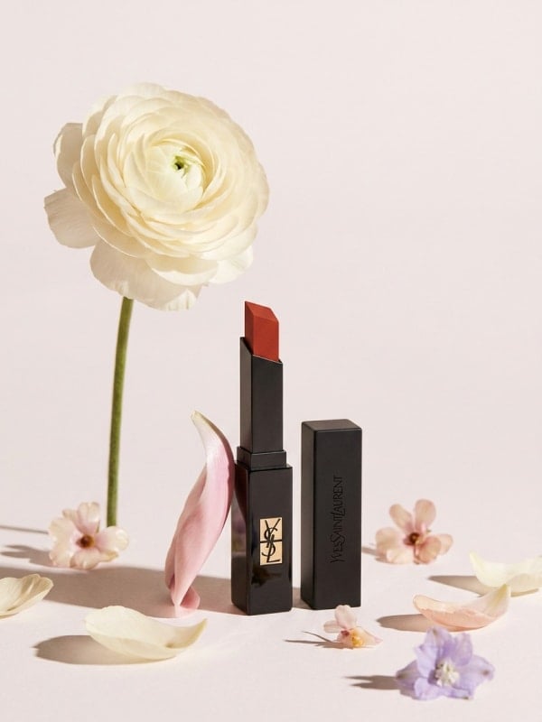 YSL slim velvet lipstick in brick red posed beside a tall white ranunculus flower and scattered pastel petals on a soft pink surface.