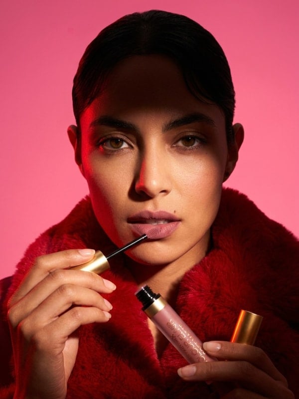 Brunette woman in a deep red faux fur coat applying Max Factor Colour Elixir liquid lipstick in a moody pink studio lit by warm red side light.