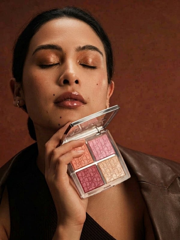 Asian woman in a brown leather blazer holding a Dior Backstage glow palette beside her face with eyes closed, in moody studio lighting on a rust-coloured backdrop.