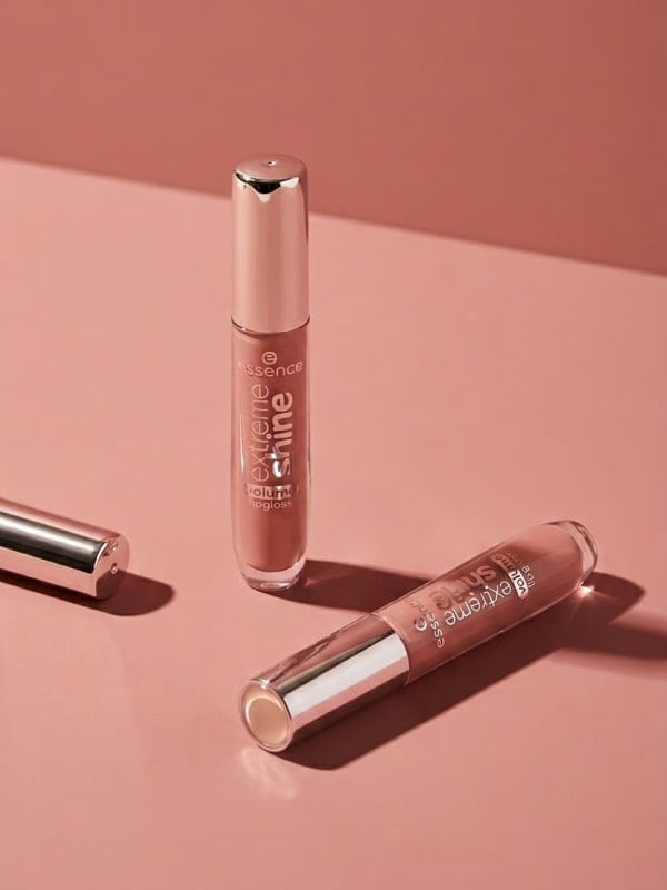 Two Essence Extreme Shine lipgloss tubes arranged as a clean still life on a layered dusty rose paper backdrop, casting soft shadows.