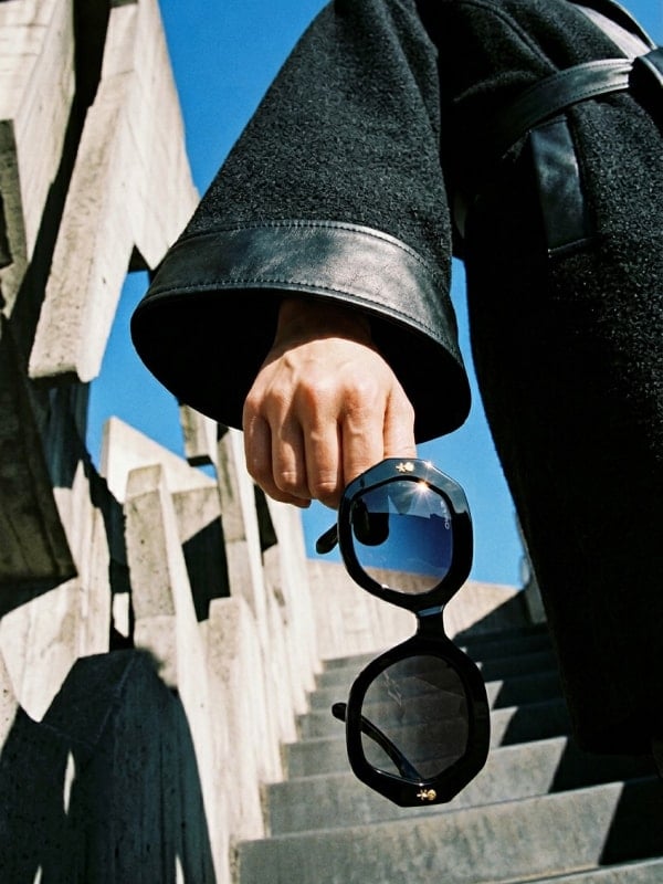 Cropped shot of a person in a black wool coat with leather cuffs holding black hexagonal sunglasses on a sunlit concrete staircase.