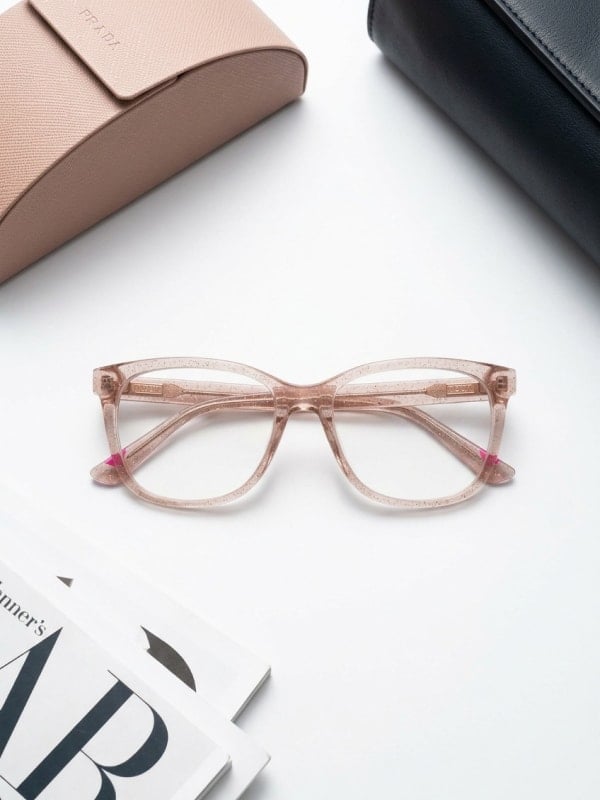 Translucent pink optical glasses laid on a white surface with a Prada glasses case and a magazine at the edges of the frame.