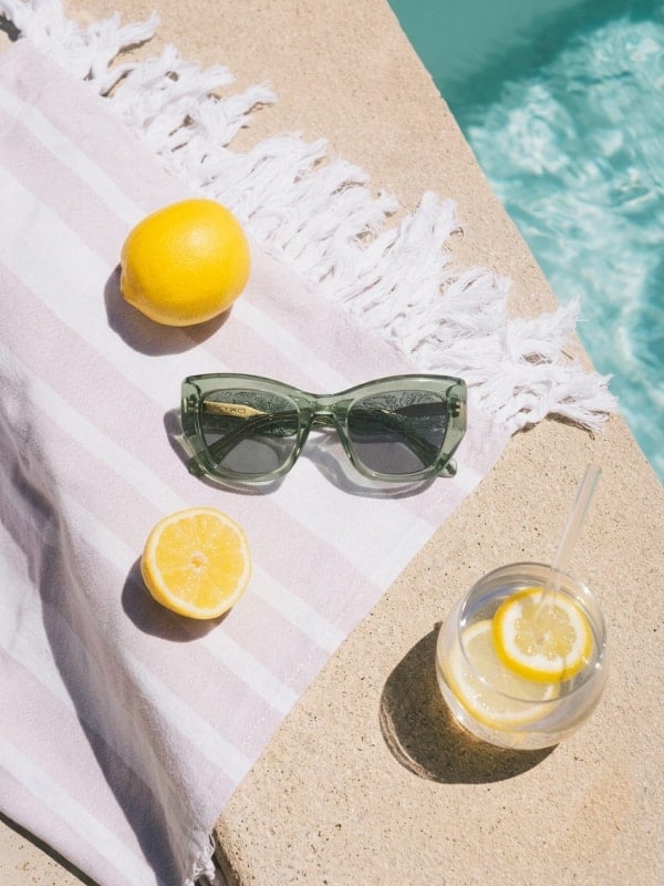Flat-lay of sage green cat-eye sunglasses on a striped beach towel beside a whole lemon, half lemon, and a glass of lemon water at the pool edge.