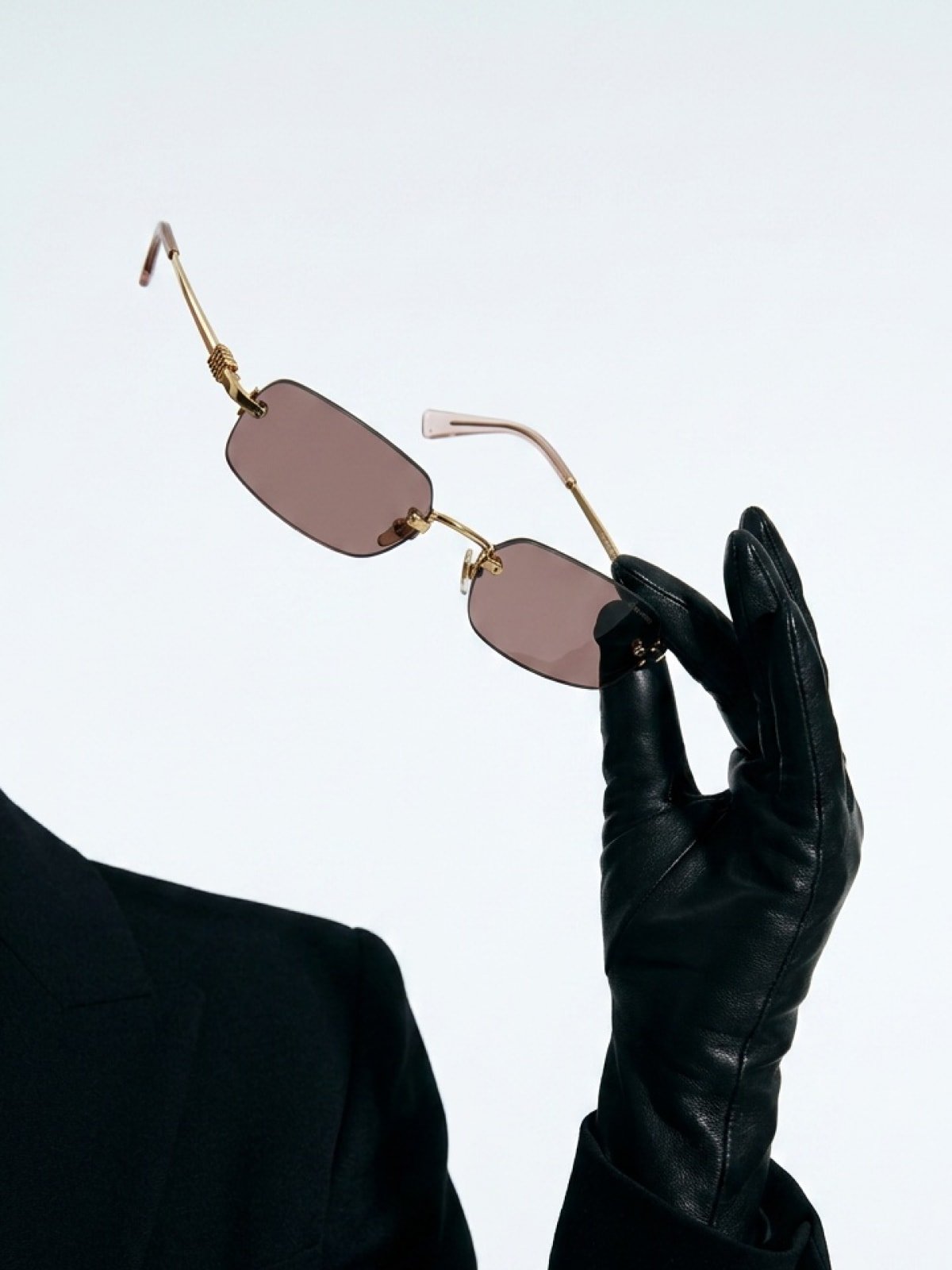 A black-leather-gloved hand holding rimless rectangular sunglasses with pink-tinted lenses, against a clean light grey background.