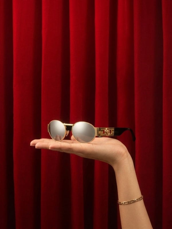 Gold oval Versace sunglasses with mirrored lenses resting on an open hand, in front of a deep red velvet curtain backdrop.