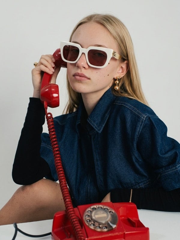 Blond woman in a denim jacket holding a red rotary phone to her ear while wearing white Etro cat-eye sunglasses, against a clean studio background.
