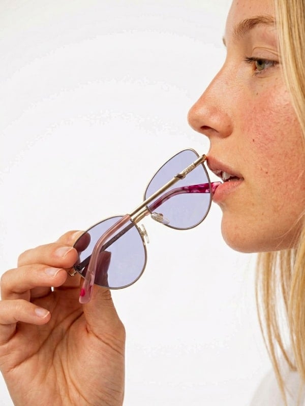 Cropped close-up of a freckled blond woman holding gold-rimmed cat-eye sunglasses with violet lenses to her lips, against a soft white background.