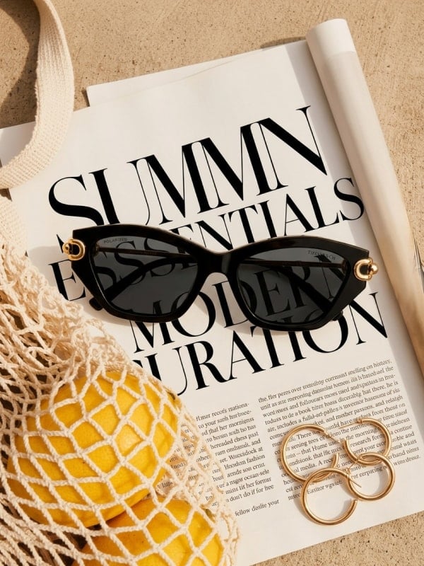 Black Tiffany cat-eye sunglasses placed on top of an open Summer Essentials magazine spread, with a mesh bag of lemons and gold rings styled around it.