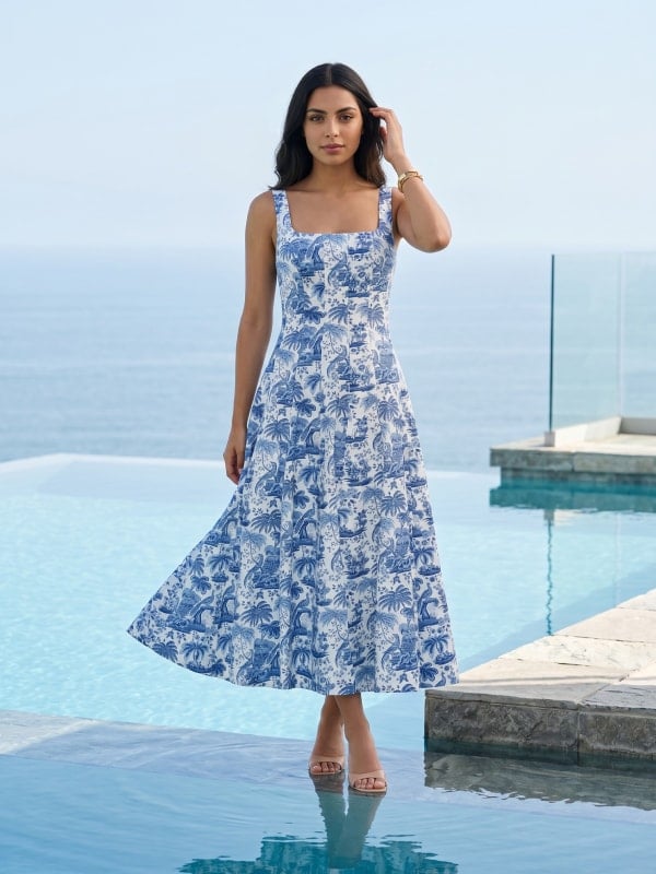 Woman in a blue and white tropical-print midi dress standing beside an infinity pool overlooking the ocean.