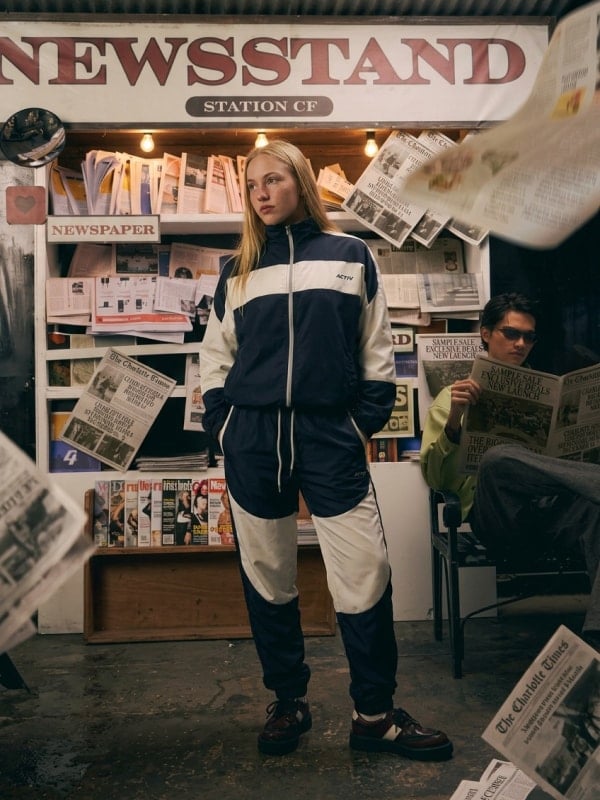 Woman wearing a navy and white Activ tracksuit standing in front of a newsstand stocked with newspapers and magazines, with a man reading in the background.