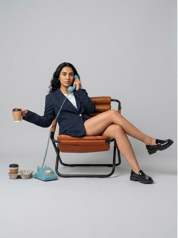 Woman in a navy pinstripe blazer seated on a leather chair holding a vintage teal rotary phone and a coffee cup, in a clean studio setting.