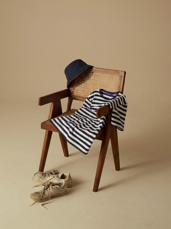 Striped Breton t-shirt draped over a wooden cane chair with a navy bucket hat resting on the backrest and worn white sneakers on the floor.