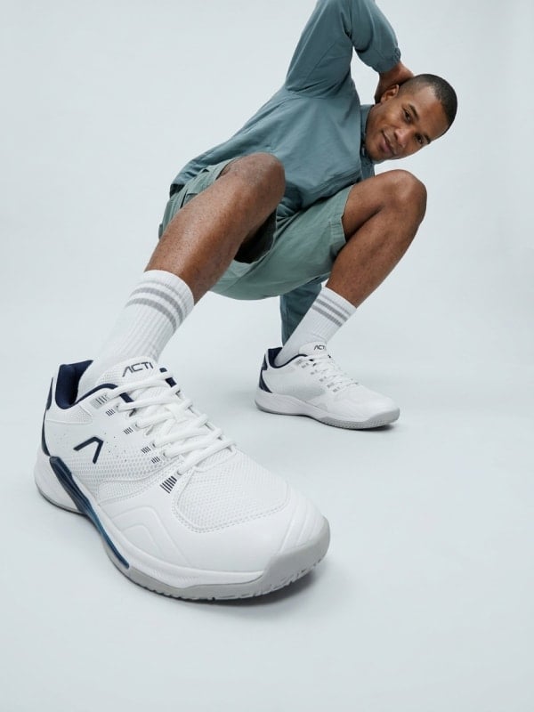 Man crouching in a clean studio wearing sage green athletic wear, white tube socks, and white Activ tennis sneakers.