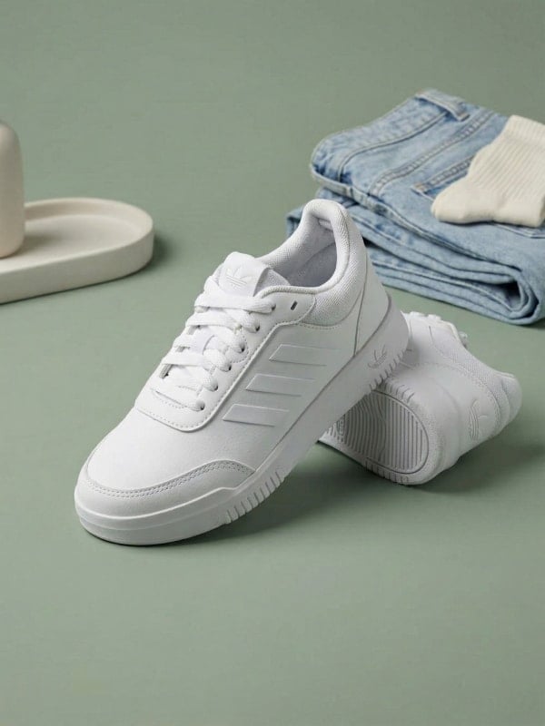 Pair of white Adidas Tensaur sneakers styled on a sage green surface with folded blue jeans and ribbed socks.