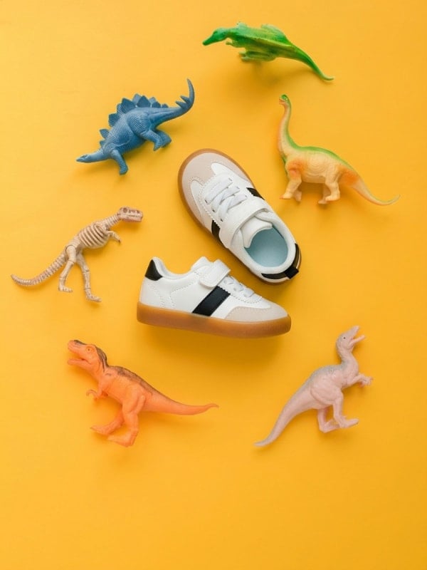 Pair of kids' white sneakers flat-lay on a yellow background, surrounded by colorful plastic toy dinosaurs.