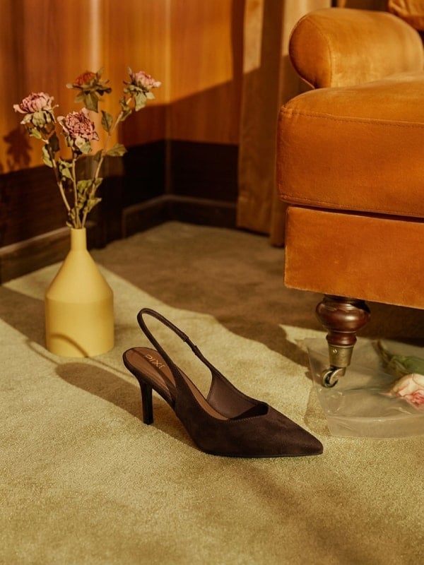 Brown suede slingback heel placed on a green carpet beside a yellow ceramic vase of dried roses, in front of a tan velvet sofa.