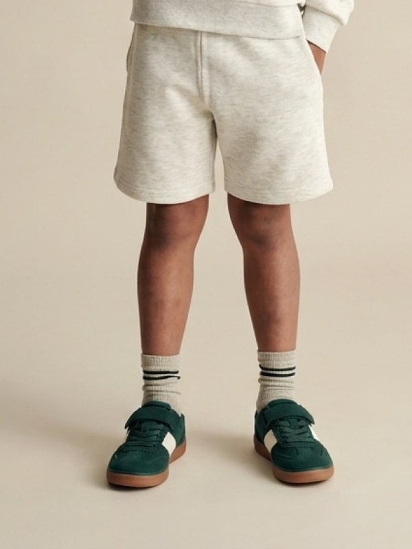 Cropped shot of a child in cream sweat shorts and ribbed socks wearing dark green velcro sneakers, against a beige studio backdrop.