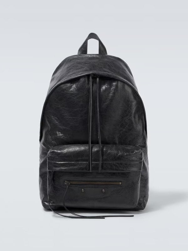Front view of a black leather backpack with a zip front pocket and long leather pull tabs, displayed on a light background.