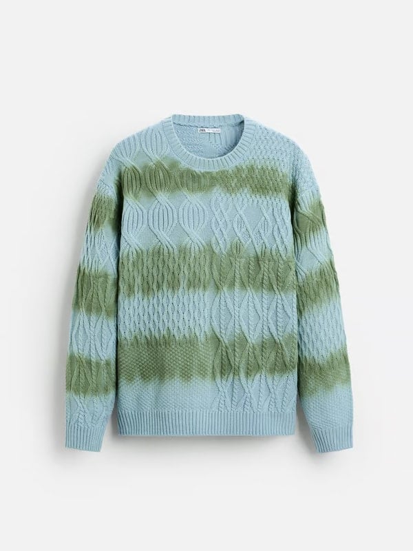 Flat lay of a light blue cable-knit sweater with green tie-dye stripes, displayed on a white background.