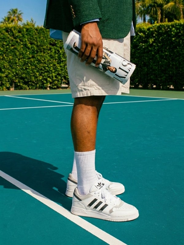 Cropped shot of a man in tan shorts and white Adidas sneakers standing on a green tennis court, holding a rolled magazine.