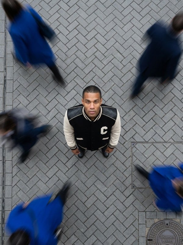 Overhead shot of a man in a black and cream Celine varsity jacket standing still on a herringbone-tiled street while blurred passersby walk around him.