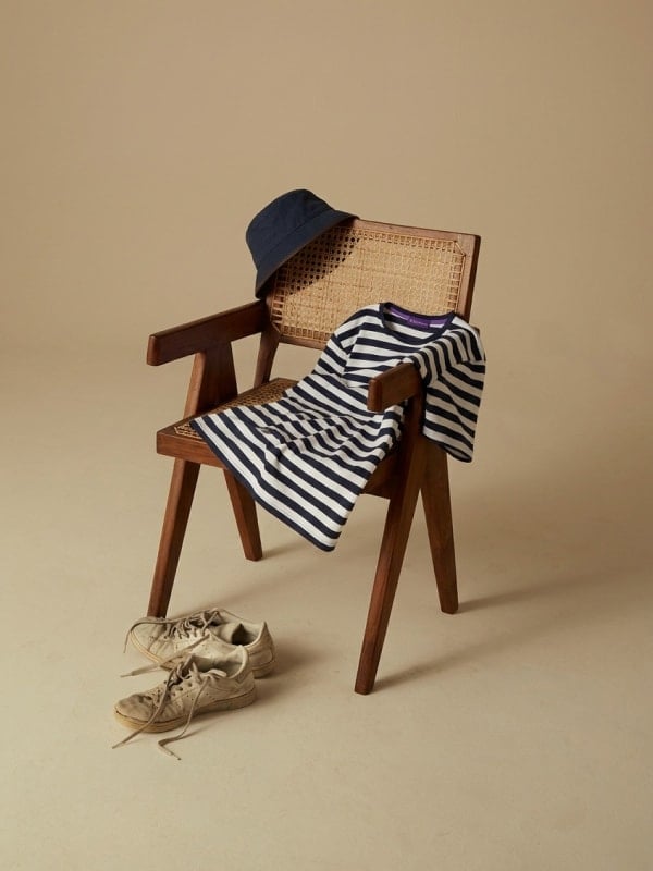 Striped Breton t-shirt draped over a wooden cane chair with a navy bucket hat resting on the backrest and worn white sneakers on the floor.