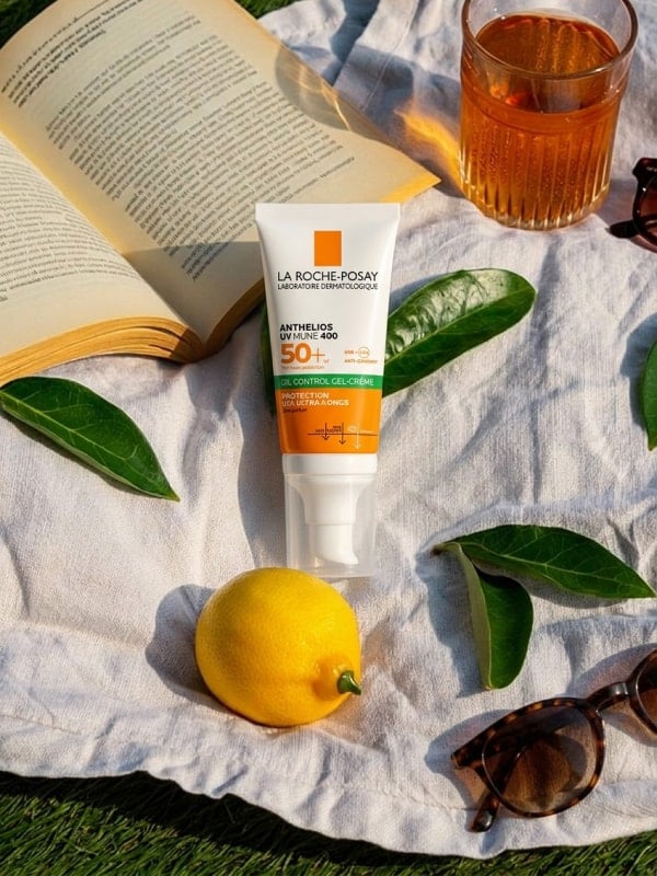 La Roche-Posay sunscreen tube laid on a white picnic cloth with a fresh lemon, leaves, an open book, sunglasses, and a glass of iced tea.