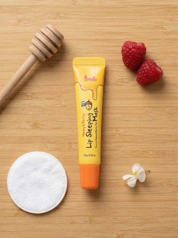 Yellow lip mask tube laid on a wooden surface beside a honey dipper, fresh raspberries, a cotton round, and a small white flower.