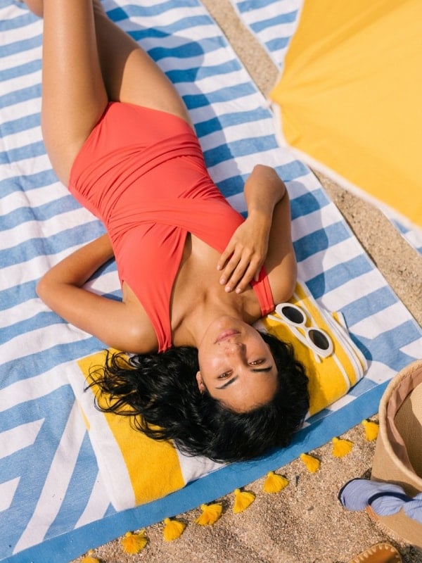 Woman in a coral red one-piece swimsuit lying on a blue and white striped beach towel in the sand, with a yellow umbrella and straw hat nearby.
