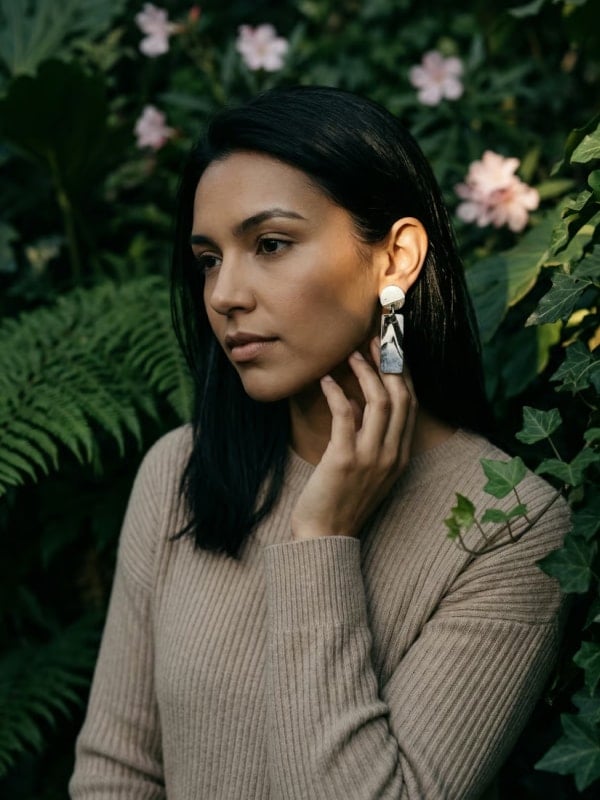 Woman with dark hair in a beige knit sweater touching her ear, wearing a white and marbled-acetate drop earring, against a backdrop of pink hibiscus flowers and ferns.