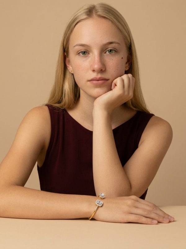 Blond woman in a sleeveless burgundy top resting her chin on her hand, wearing a gold open-cuff diamond bangle, against a warm tan studio background.