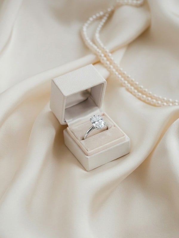 Silver solitaire engagement ring presented in a cream velvet ring box on rippled ivory satin, with a strand of pearls draped behind it.