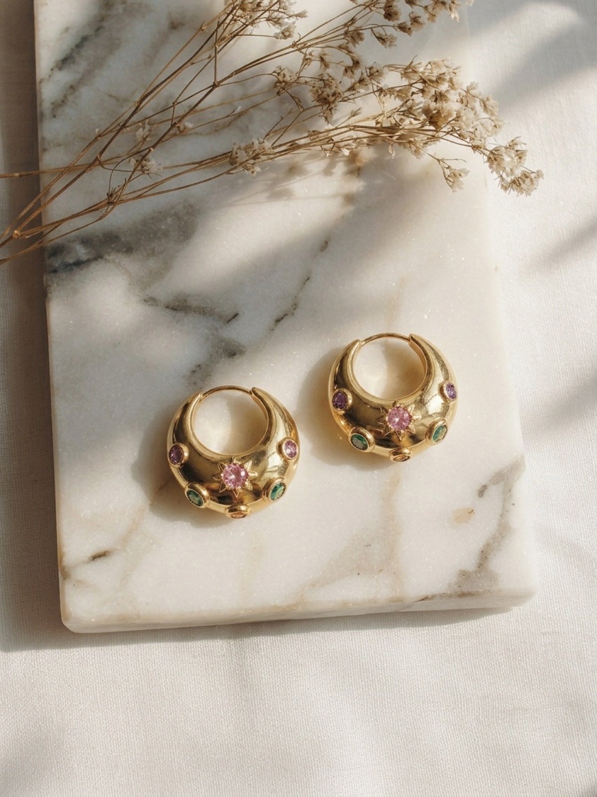 Pair of chunky gold gemstone hoop earrings laid on a marble slab with sprigs of dried flowers casting soft shadows in warm sunlight.
