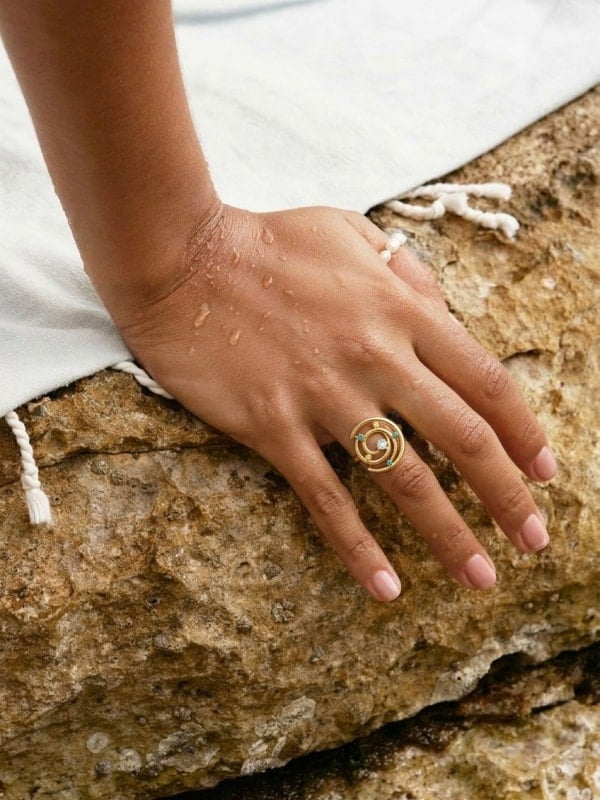 A wet hand wearing a gold turquoise spiral ring resting on textured beige rock at the edge of a poolside towel.