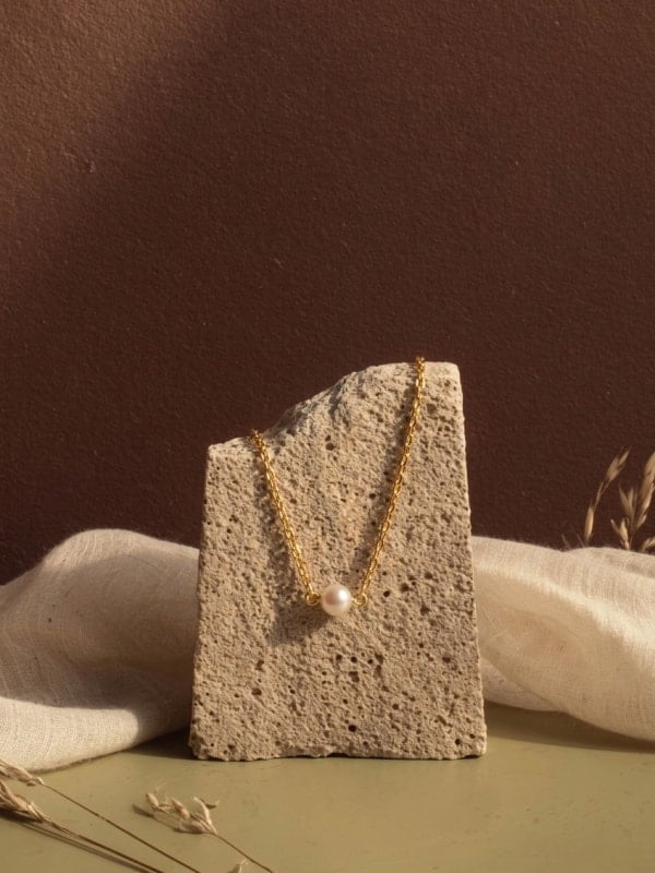 Gold chain necklace with a pearl pendant draped over a textured stone block, styled on a green surface with linen fabric and dried grasses.