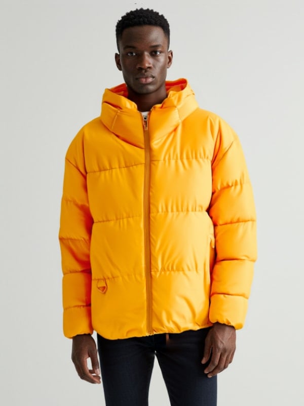 Man wearing a bright yellow puffer jacket with a hood, paired with dark jeans, standing against a plain background.