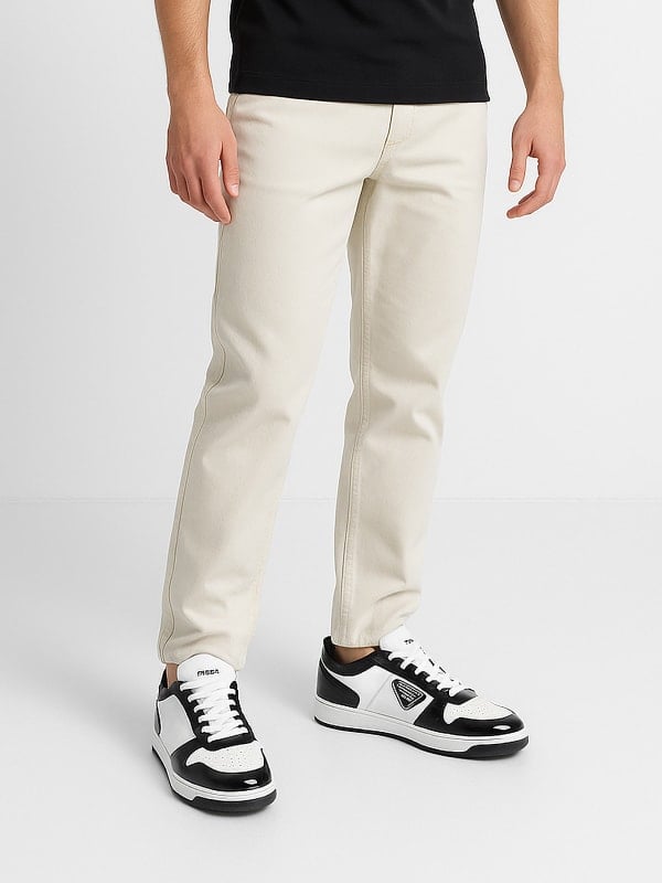 Man wearing off-white jeans and black-and-white sneakers, paired with a black top, standing against a minimal white background.