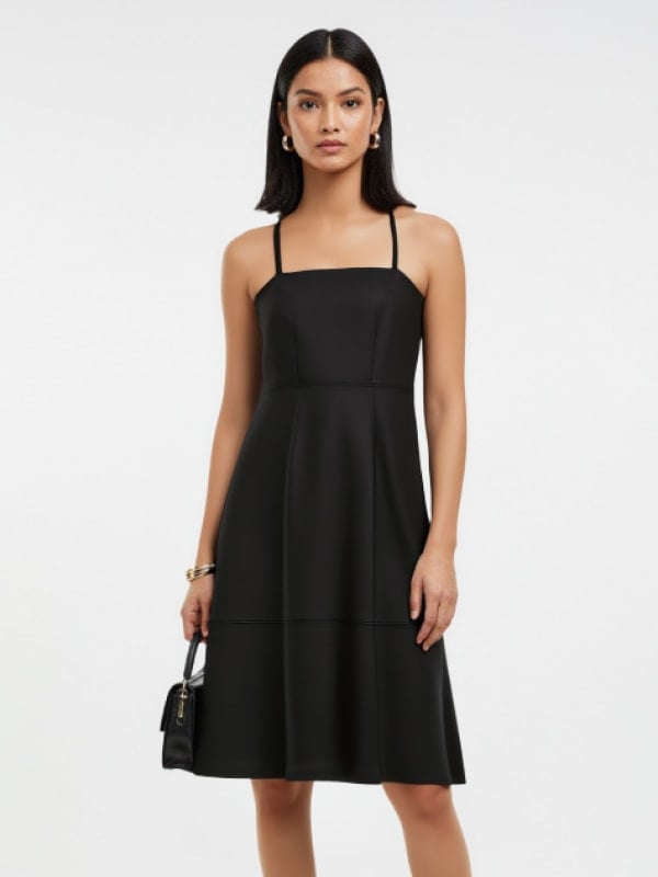 Woman in a sleek black midi dress with thin straps, accessorized with a small black handbag and gold jewelry, standing against a plain background.