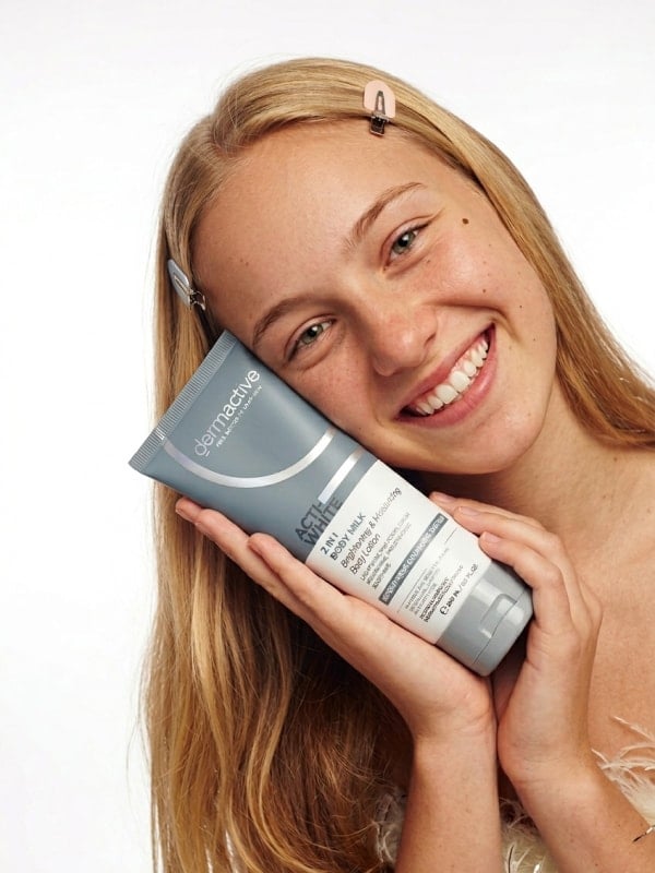 Smiling young woman with long blond hair holding a Dermactive Acti-White body milk tube against her cheek on a clean studio background.