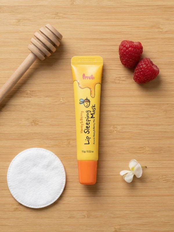 Yellow lip mask tube laid on a wooden surface beside a honey dipper, fresh raspberries, a cotton round, and a small white flower.